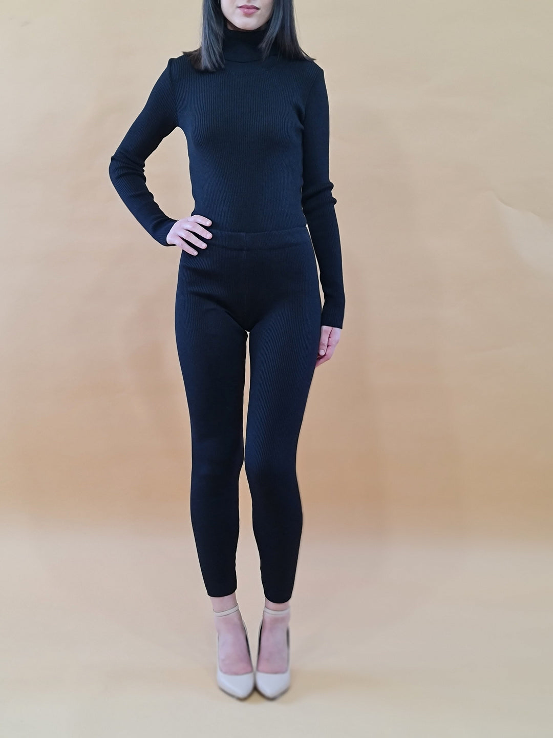 Woman in black turtleneck sweater and leggings, beige heels, standing against beige background