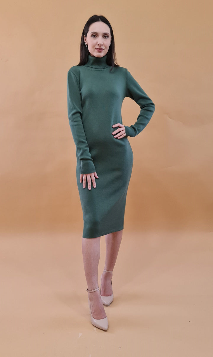 Woman in a green long-sleeve dress standing with one hand on hip against a beige background