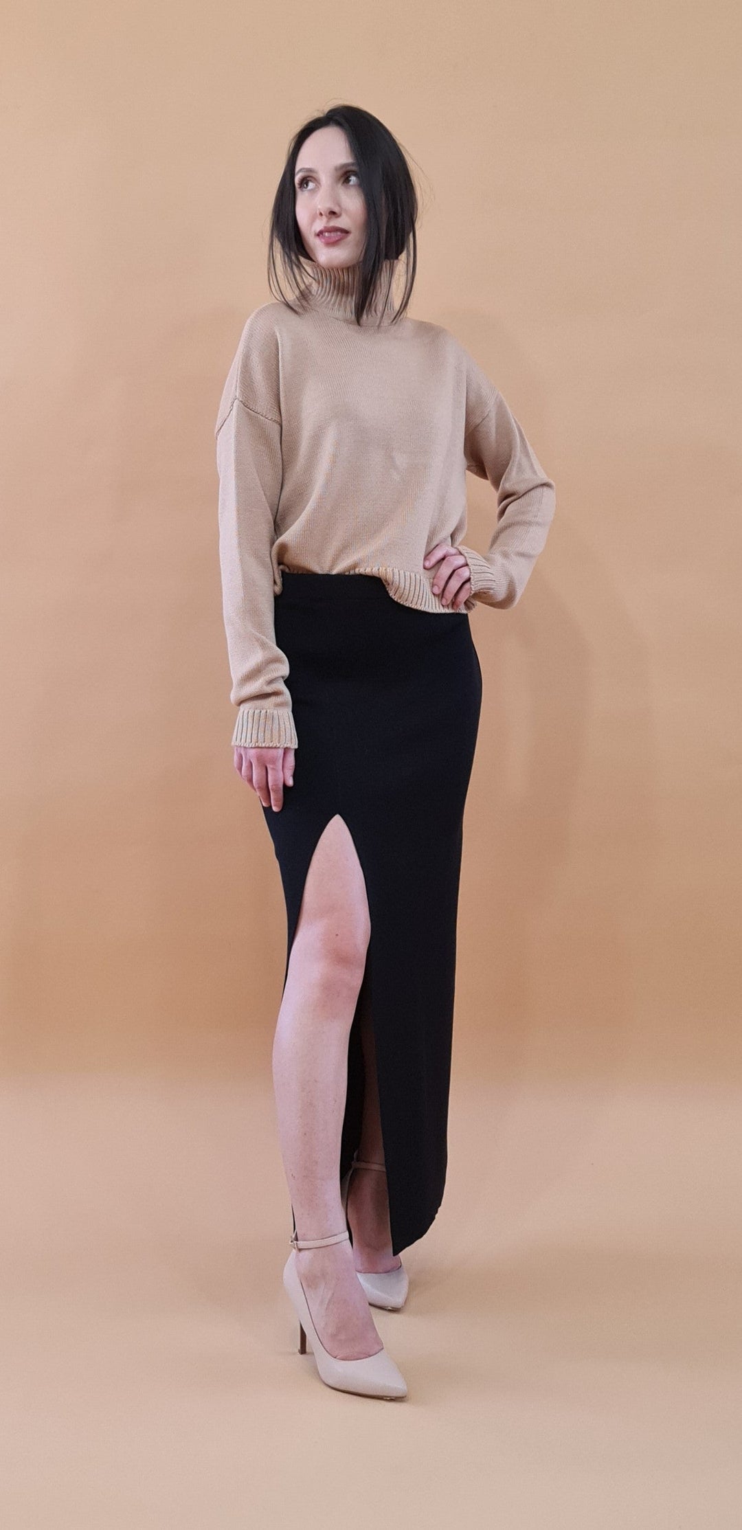 Woman wearing a beige turtleneck sweater and black skirt with a high slit, standing in front of a beige background
