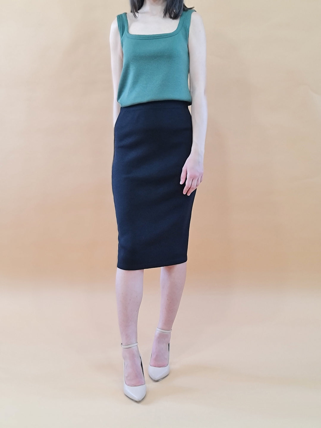 Woman wearing green sleeveless top and black pencil skirt with high heels against tan background