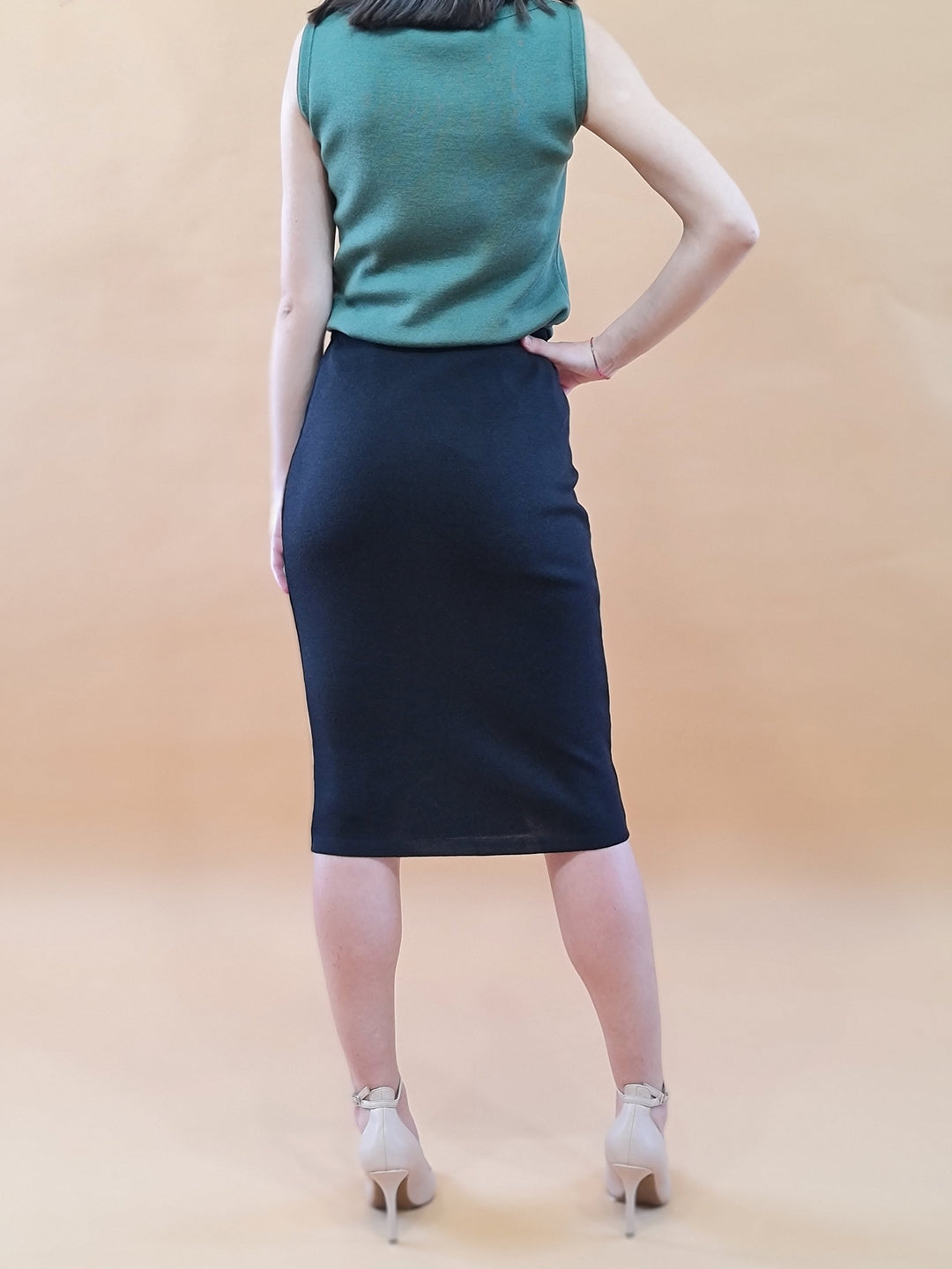 Woman wearing green sleeveless top with black pencil skirt and beige heels, standing on light brown background