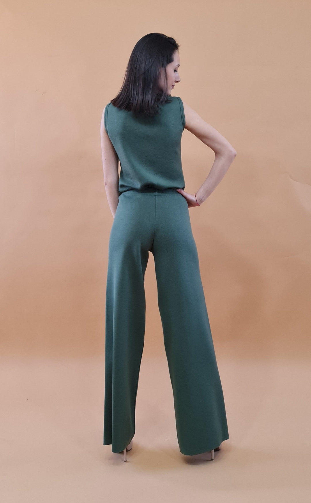 Woman wearing green sleeveless top and wide-leg pants, standing against beige background with back facing camera