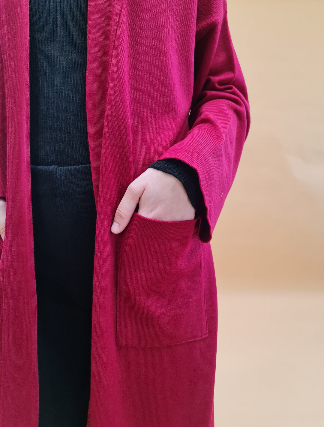 Close-up of a person wearing a long, red cardigan with their hands in the pockets against a neutral background