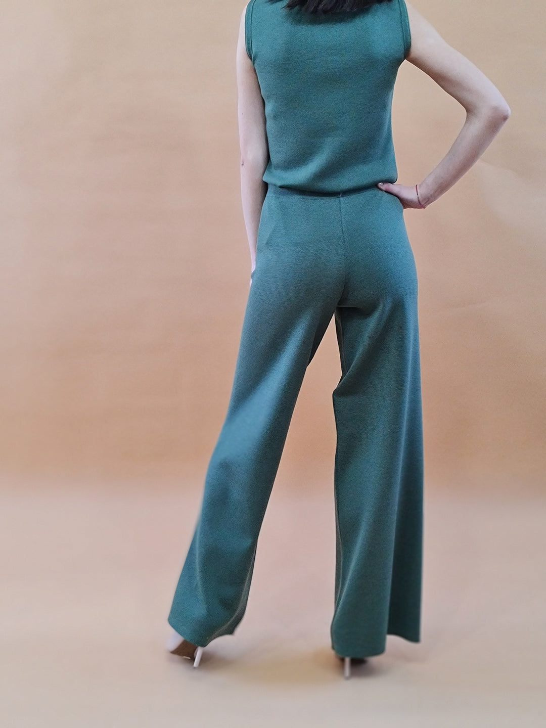 Woman in stylish green sleeveless top and wide-leg pants standing against a plain background