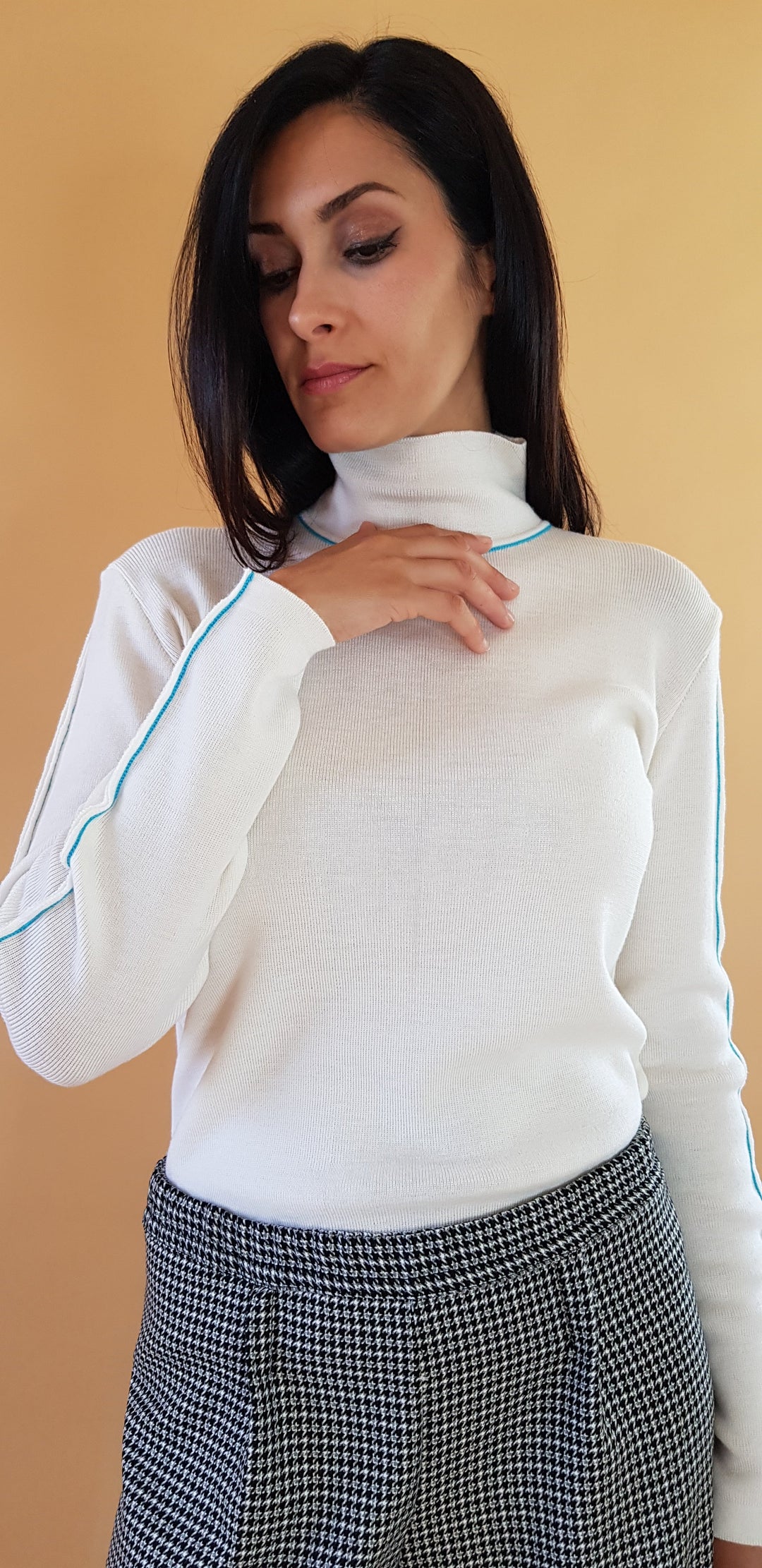 Woman wearing a white turtleneck sweater with teal accents and checked pants against an orange background