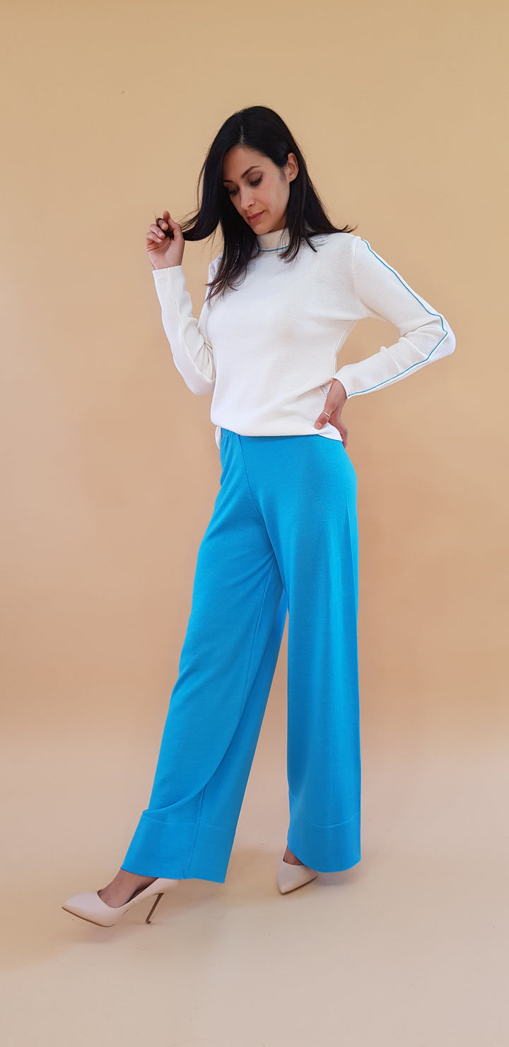 Woman wearing white sweater with blue wide-leg trousers and high heels posing confidently against beige background