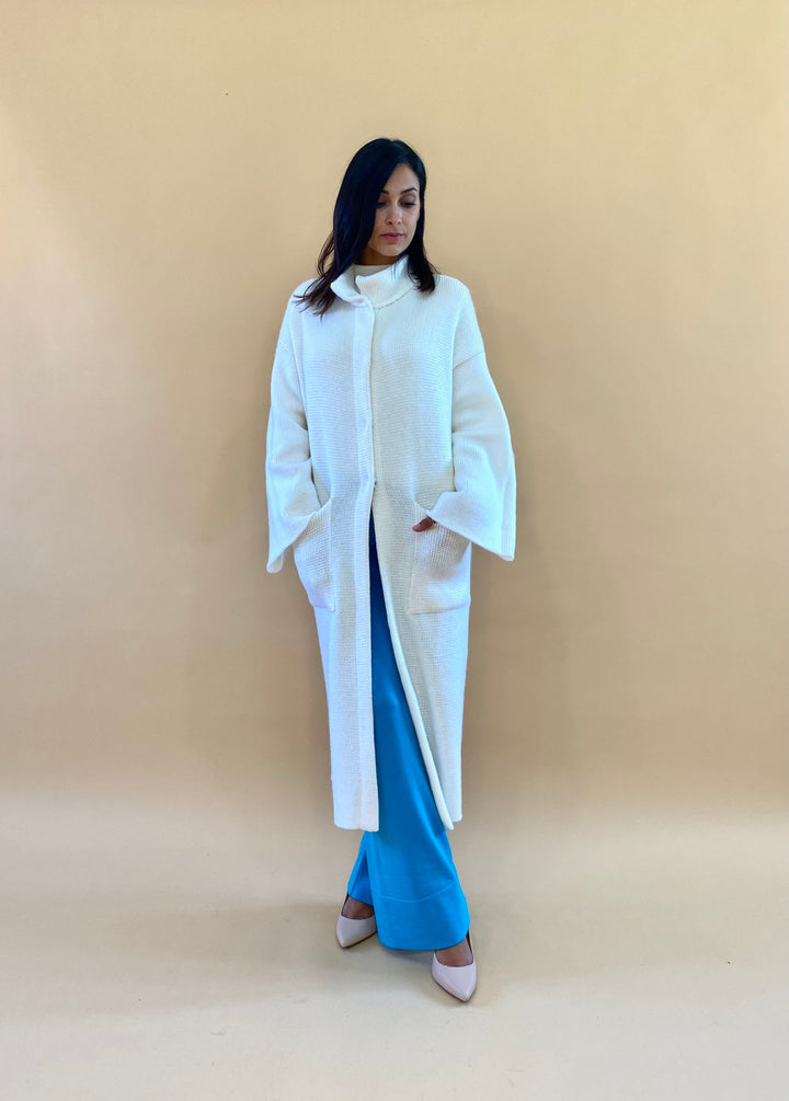 Woman in white long coat and blue pants standing against a beige background