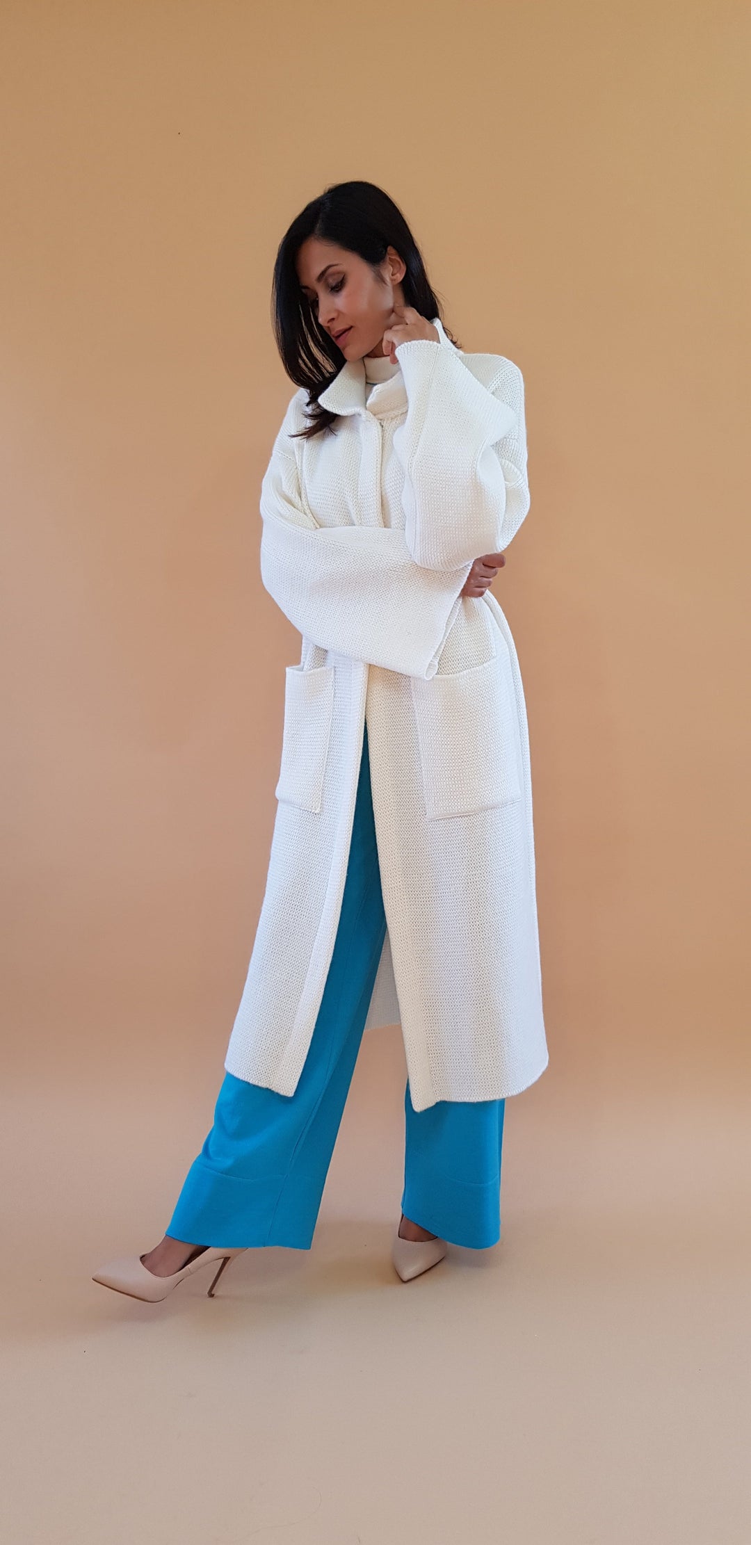 Woman wearing a white oversized coat with blue pants and nude heels against a beige background