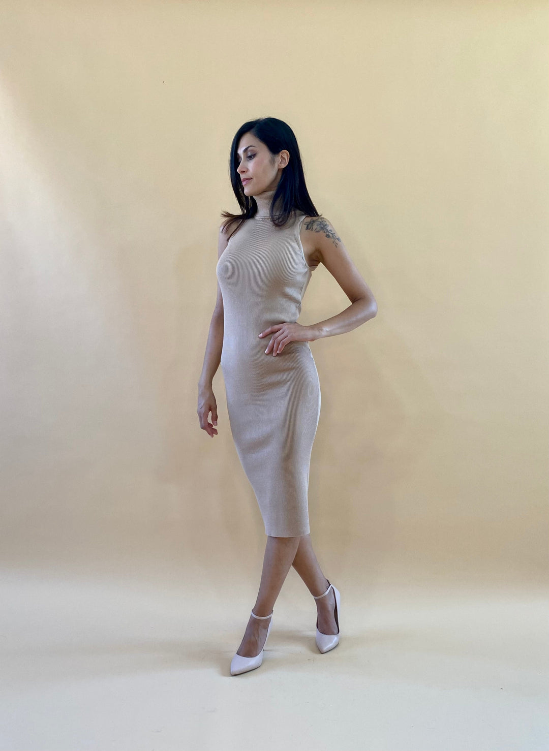 Woman wearing a fitted beige sleeveless dress and white heels against a beige background