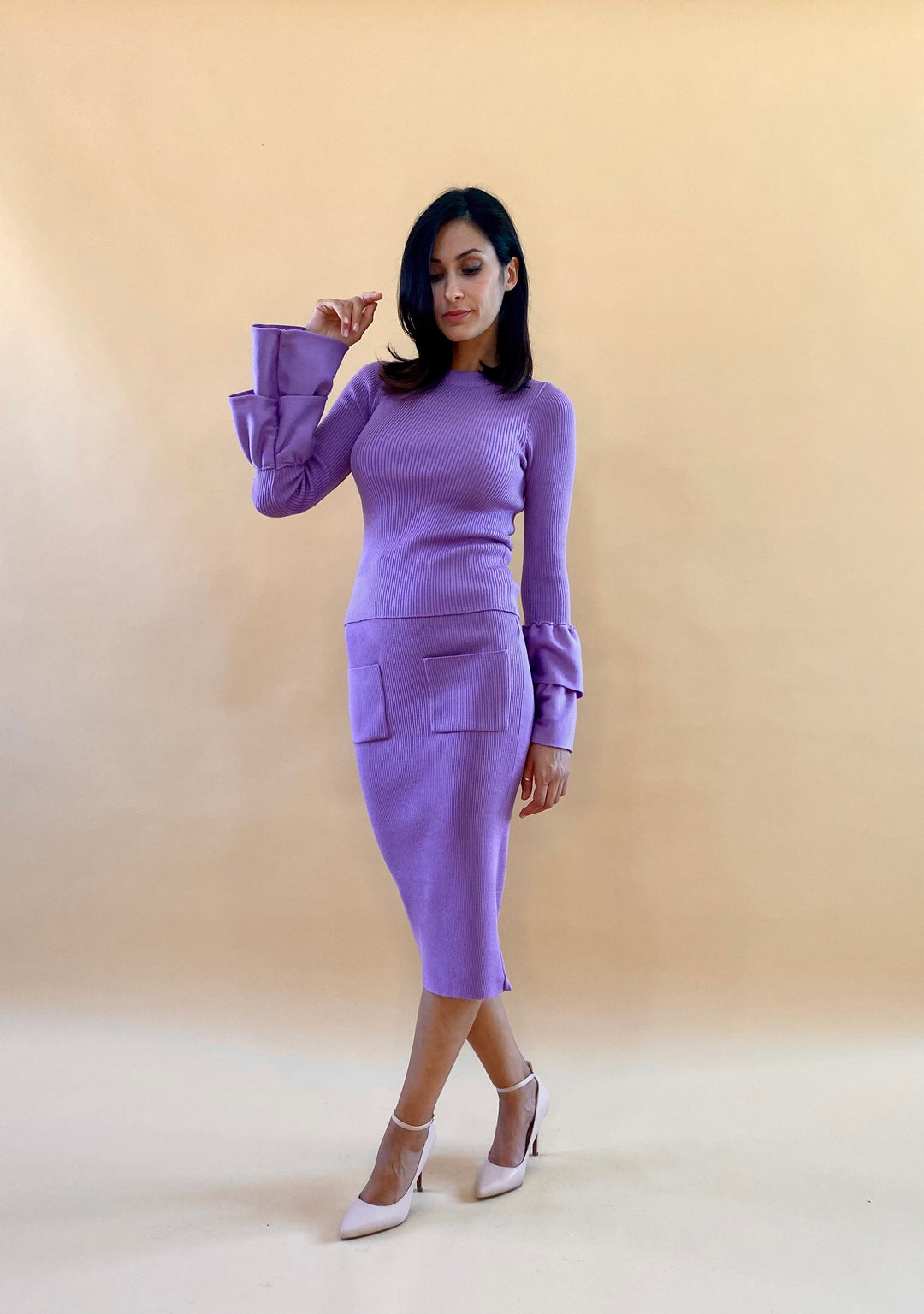 Woman wearing a fitted purple outfit with long sleeves and matching pencil skirt, posing against a beige background