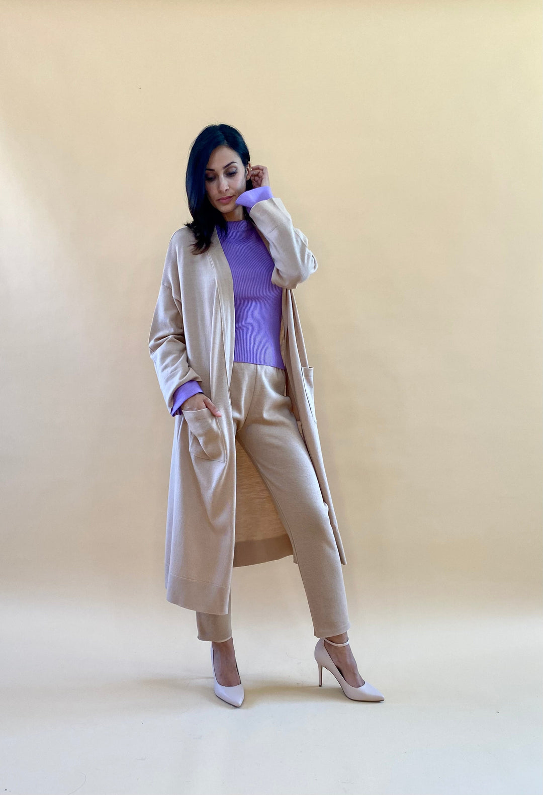 Woman wearing beige coat, matching pants, purple sweater, and heels against beige background