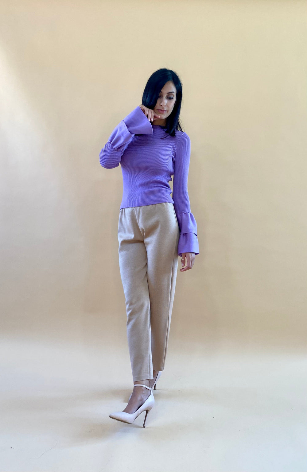 Woman wearing a purple long-sleeve top with bell sleeves and beige pants posing against a beige background