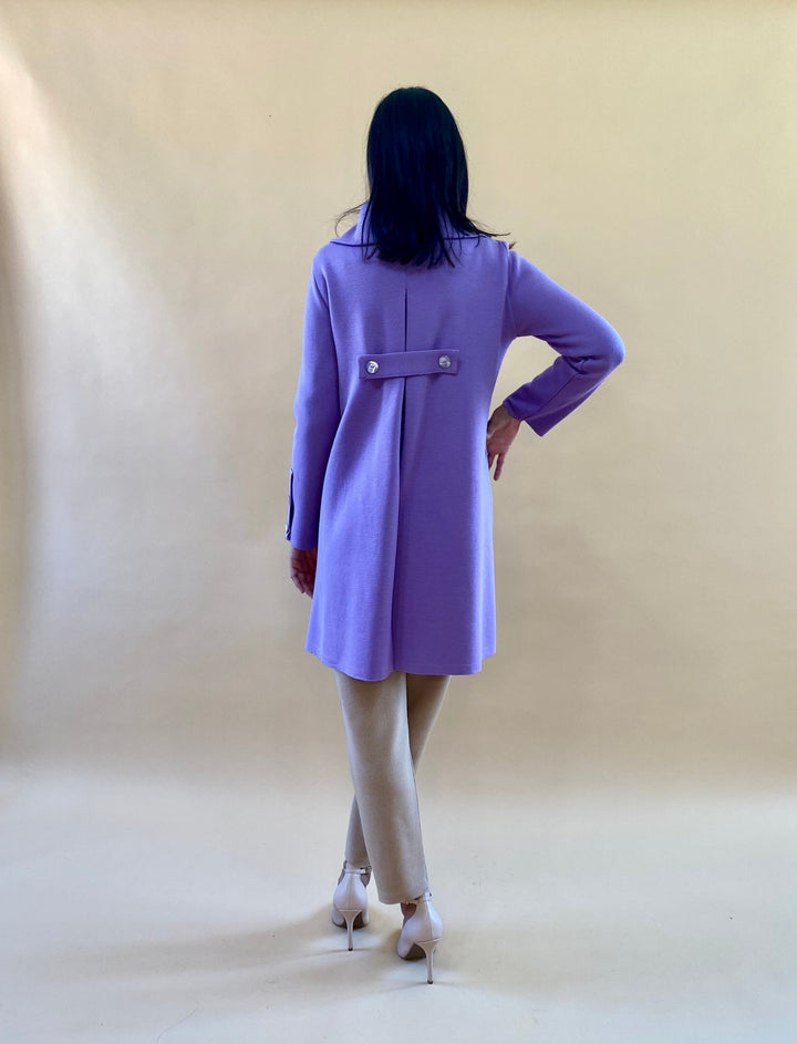 Woman wearing a lavender coat and beige pants standing against a light beige background, facing away