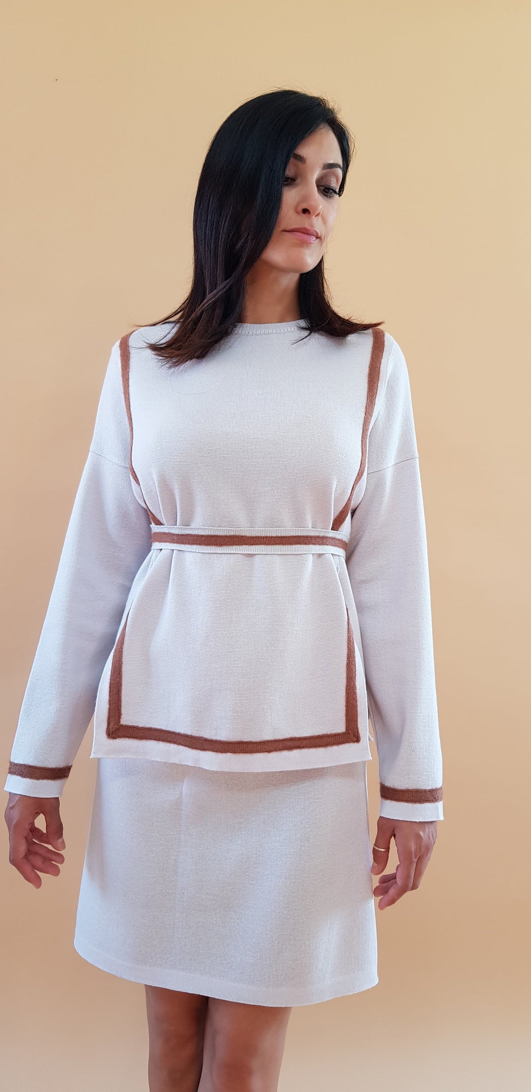 Woman wearing a white sweater and skirt set with brown trim, standing against a beige background