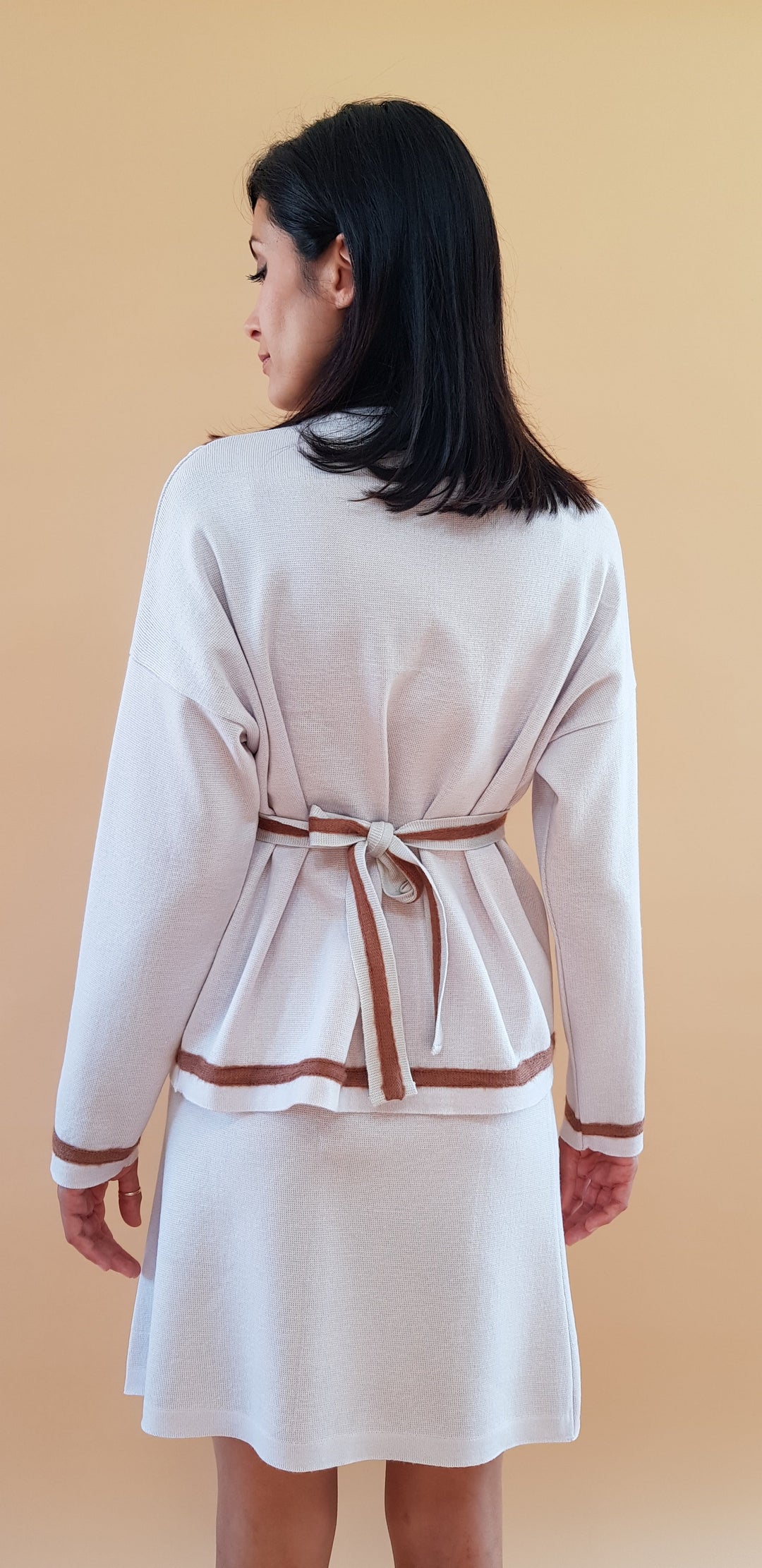 Woman wearing beige belted cardigan and dress set standing against a peach background