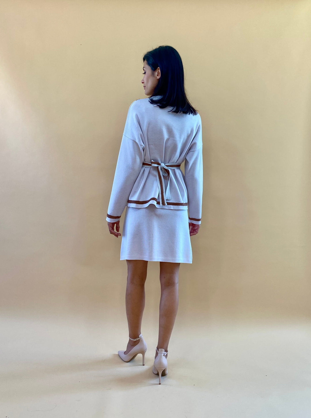 Woman wearing a white dress with a belt, viewed from the back, standing in front of a beige background