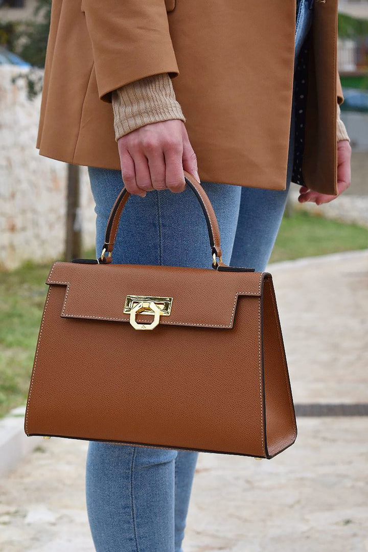 Person holding a stylish brown leather handbag with gold clasp and handle, wearing blue jeans and tan coat