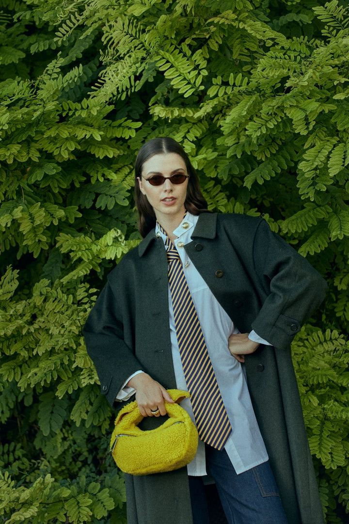 Woman in stylish outfit with striped tie and yellow handbag standing in front of leafy green background