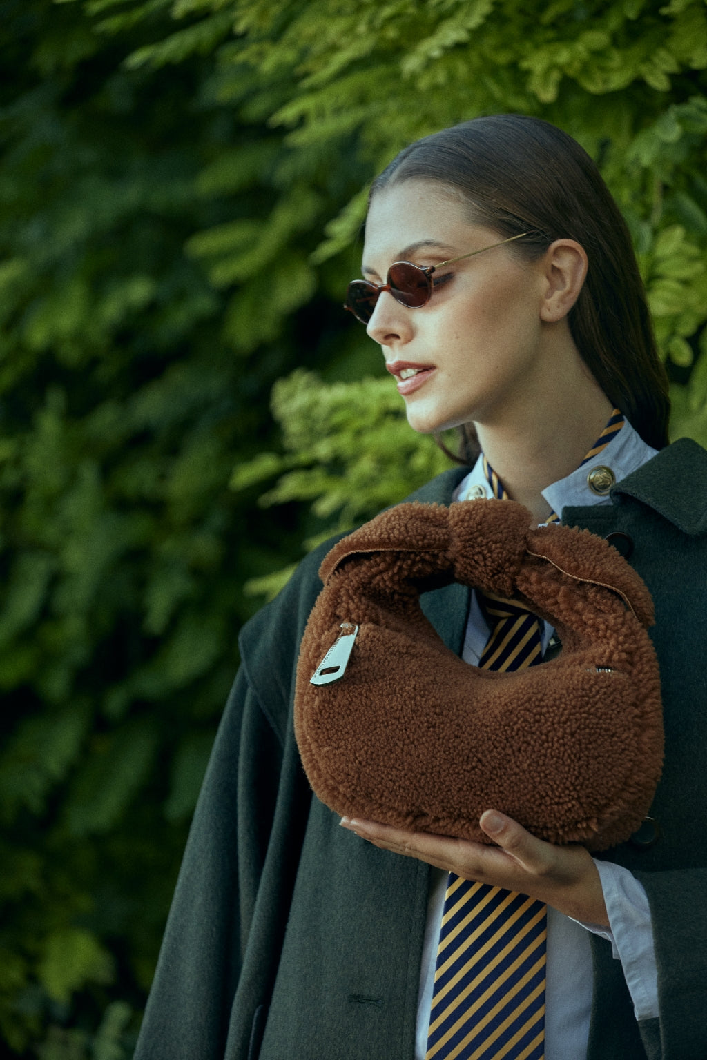 Person wearing sunglasses holding a brown plush handbag against a leafy green background