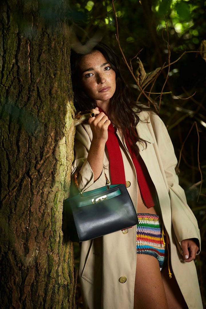 Woman in beige coat and red scarf leaning against tree in forest, holding black handbag, wearing colorful crochet shorts