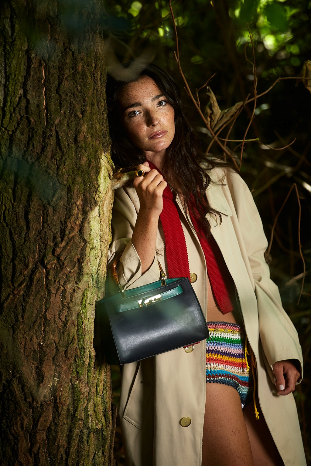 Woman in a forest wearing a trench coat, holding a black handbag, and colorful knitted shorts