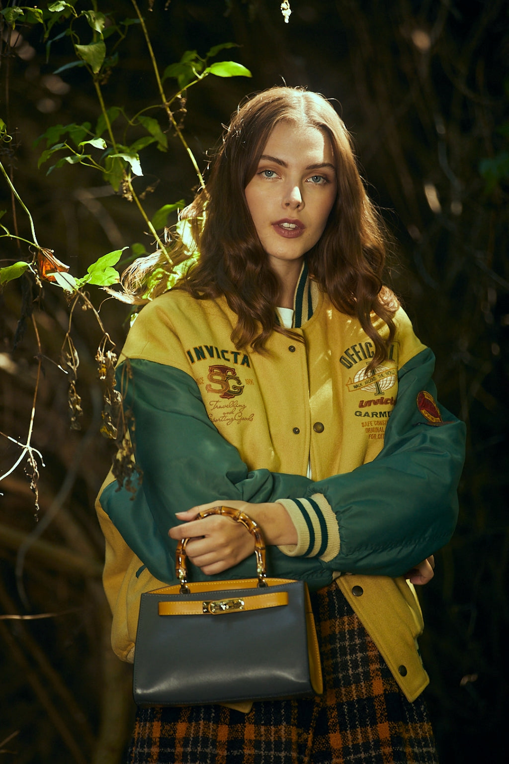 Young woman in a yellow varsity jacket holding a stylish handbag in a forest setting