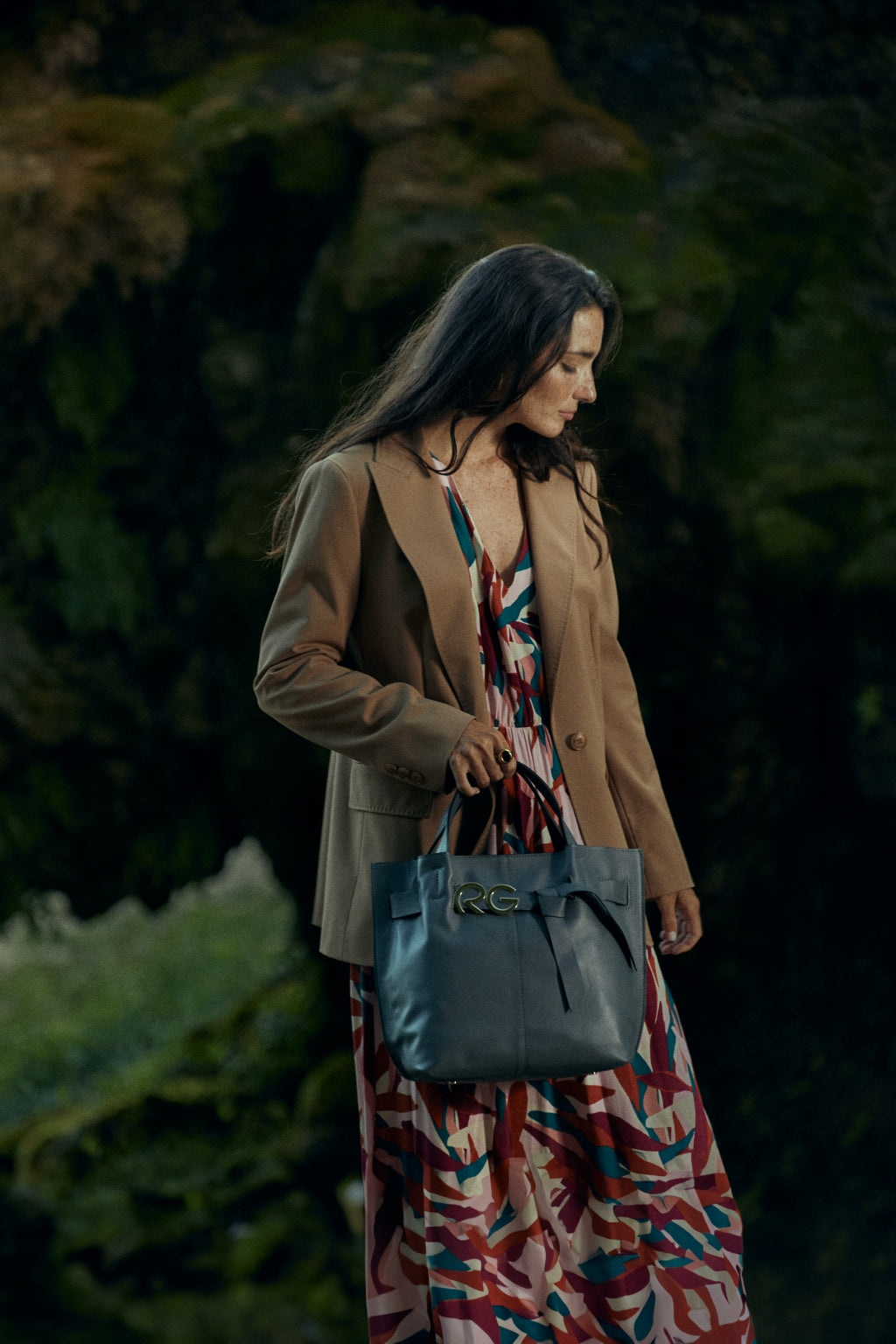 Woman in colorful dress and brown blazer holding a designer handbag outdoors