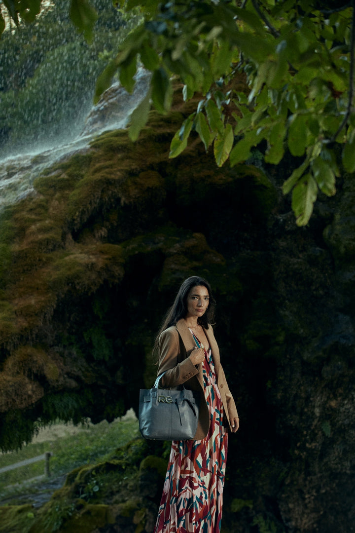 Woman in a colorful dress and coat holding a gray handbag standing in a lush, green forest