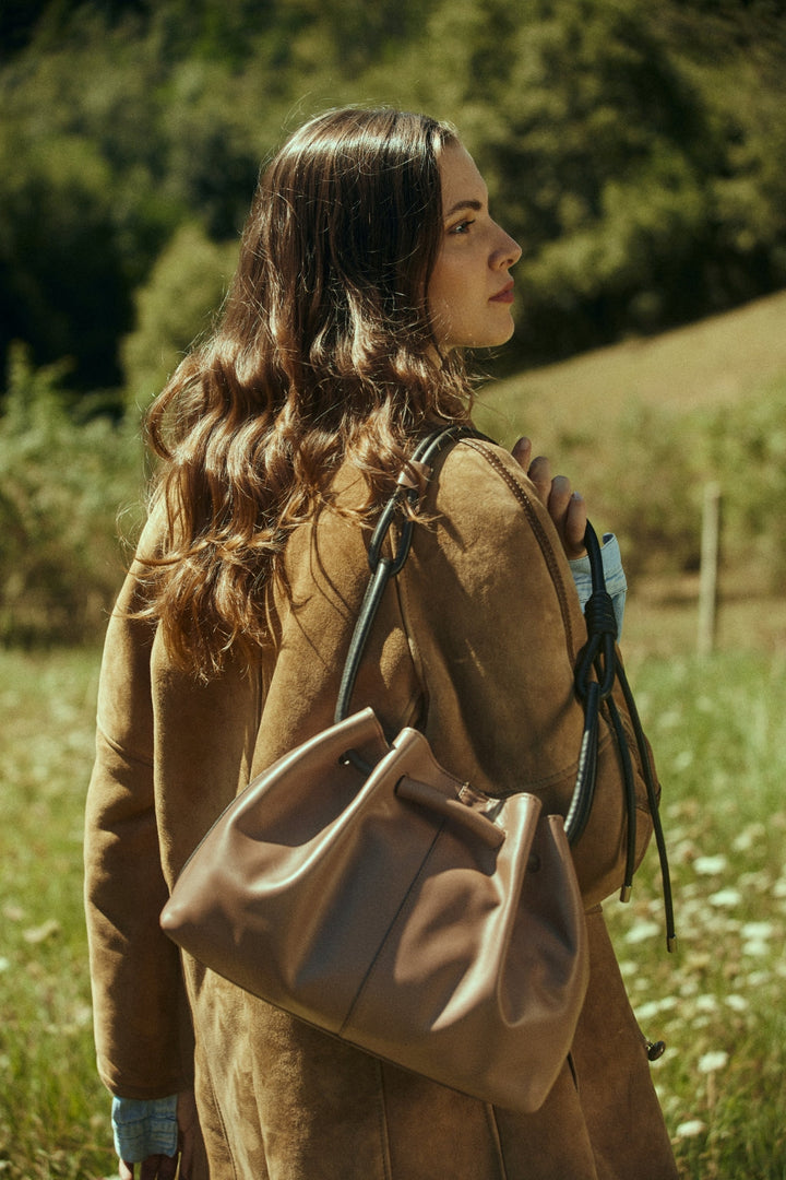 Woman standing in a field wearing a brown coat and holding a leather handbag