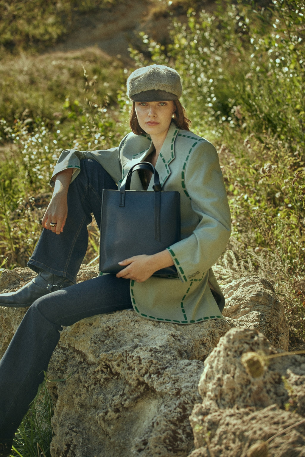 Woman wearing green blazer and cap sitting on rock holding a black handbag