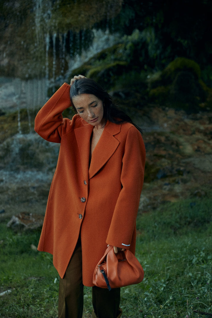 Woman in an orange coat holding a matching purse near a waterfall