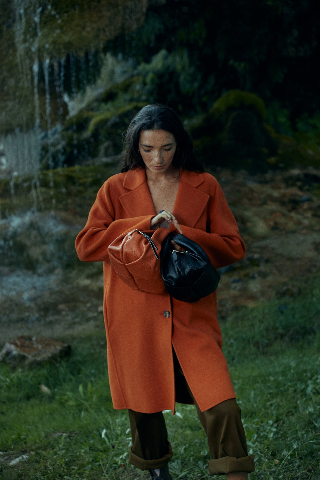 Woman in an orange coat holding two purses standing outdoors near a waterfall