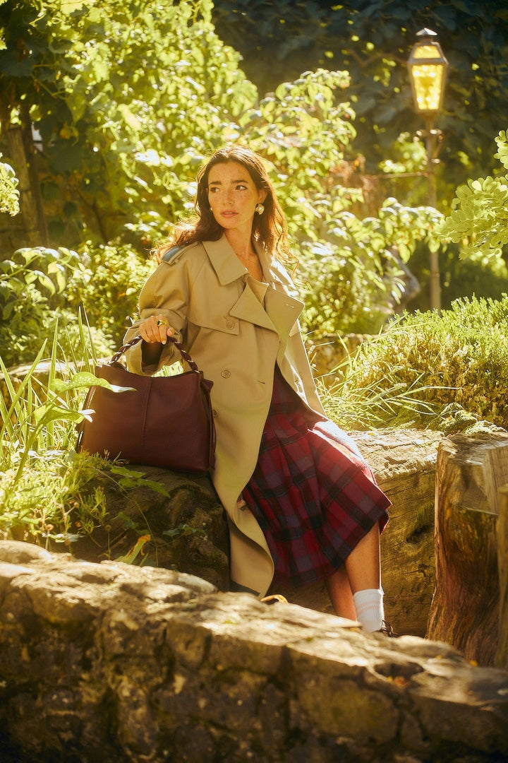 Woman in a stylish trench coat and plaid skirt sitting outdoors with a leather bag in a lush garden