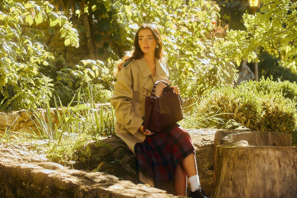 Woman sitting in a lush garden holding a handbag, wearing a beige trench coat and a plaid skirt