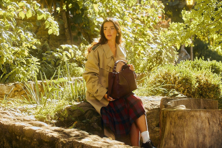 Woman sitting in a lush garden holding a handbag, wearing a beige trench coat and a plaid skirt