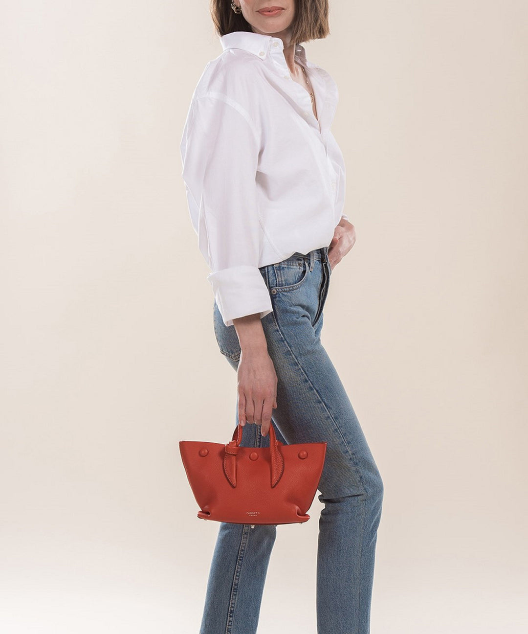 Woman in white shirt and blue jeans holding small red handbag against beige background