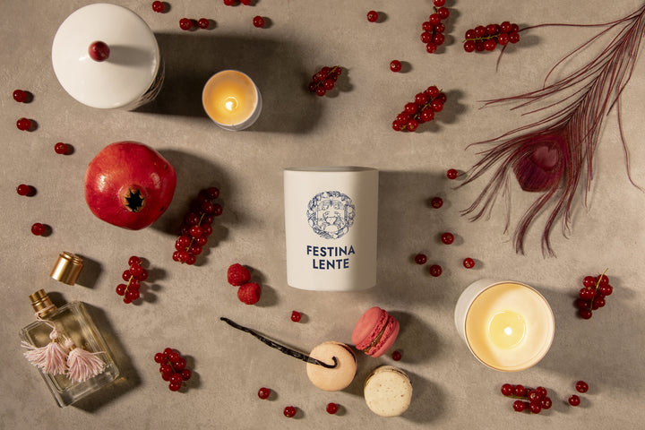 Flat lay of Festina Lente candle surrounded by fruits, macarons, candles, and perfume bottle on a textured surface