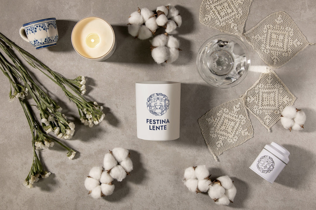 Festina Lente candle with cotton flowers, cup of tea, glass of water, and delicate lace on a textured surface