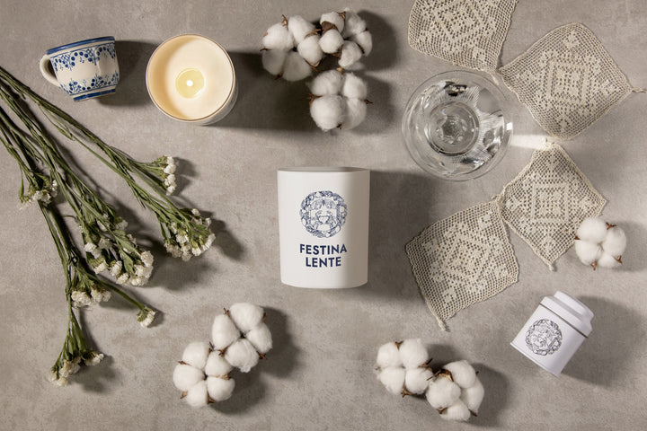 Festina Lente candle with cotton flowers, cup of tea, glass of water, and delicate lace on a textured surface