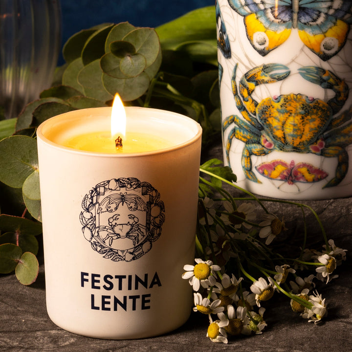 Lit Festina Lente candle surrounded by flowers and greenery with a decorative cup in the background