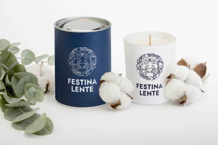 Festina Lente candle in navy blue tin and white jar surrounded by cotton and eucalyptus on white background