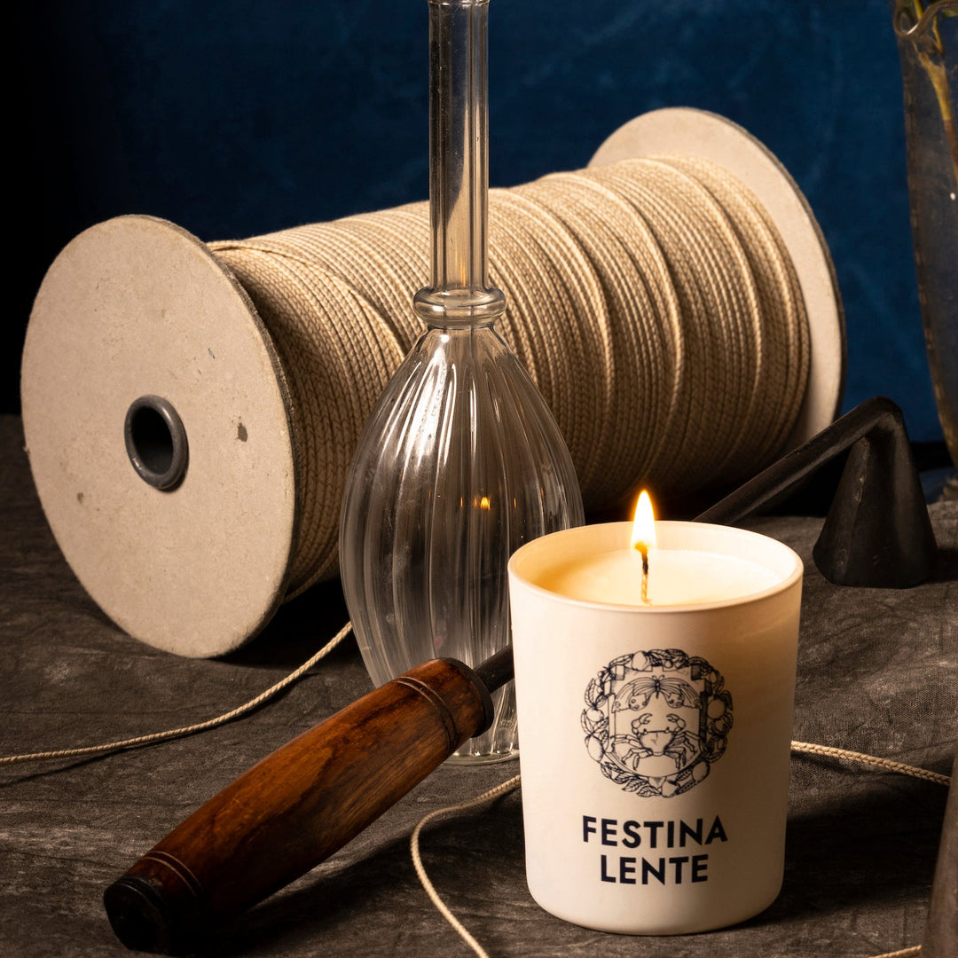 Lit Festina Lente candle placed beside crafting tools on a dark surface