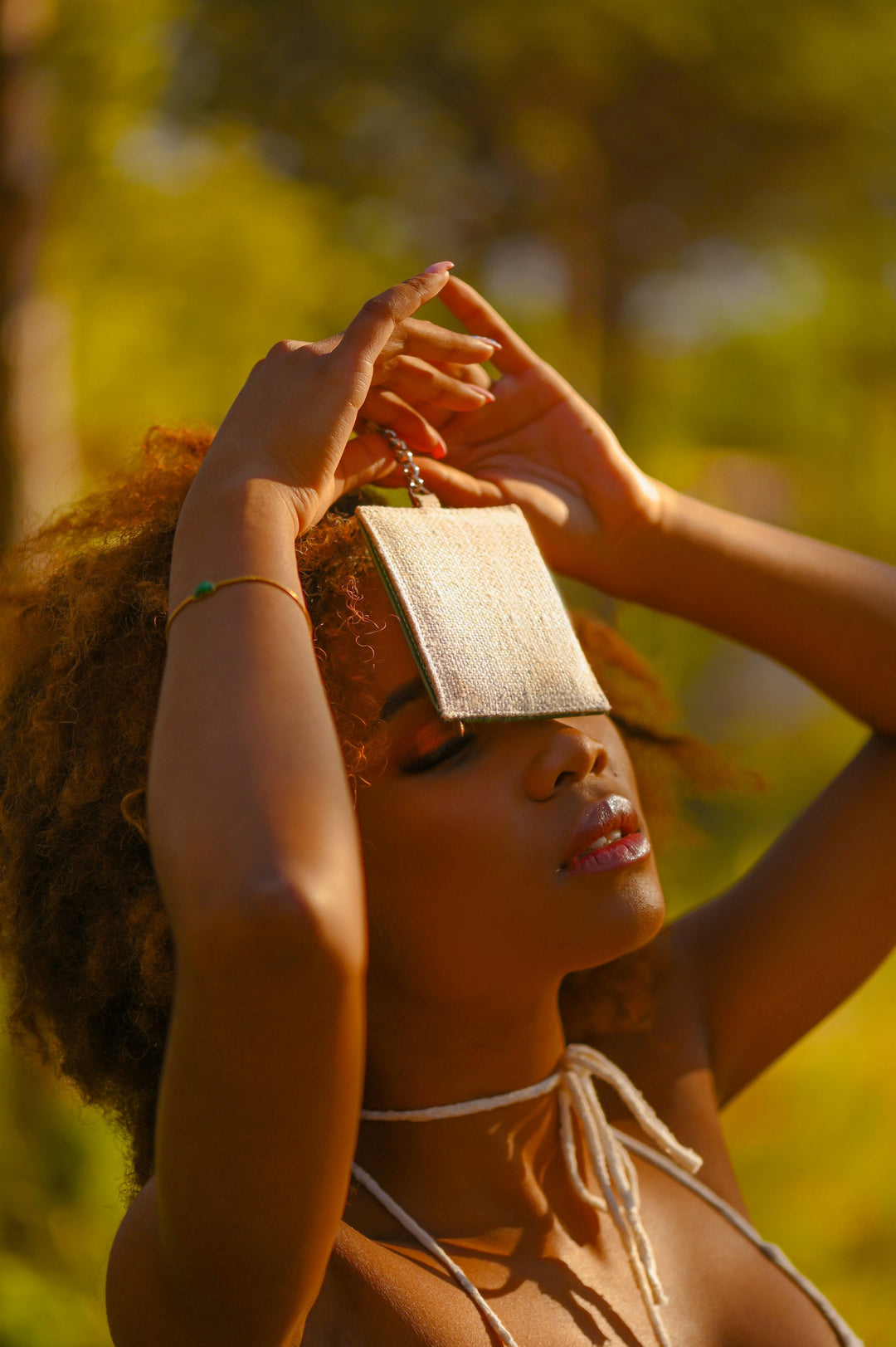 Woman with curly hair posing outdoors, holding a small silver purse to her forehead