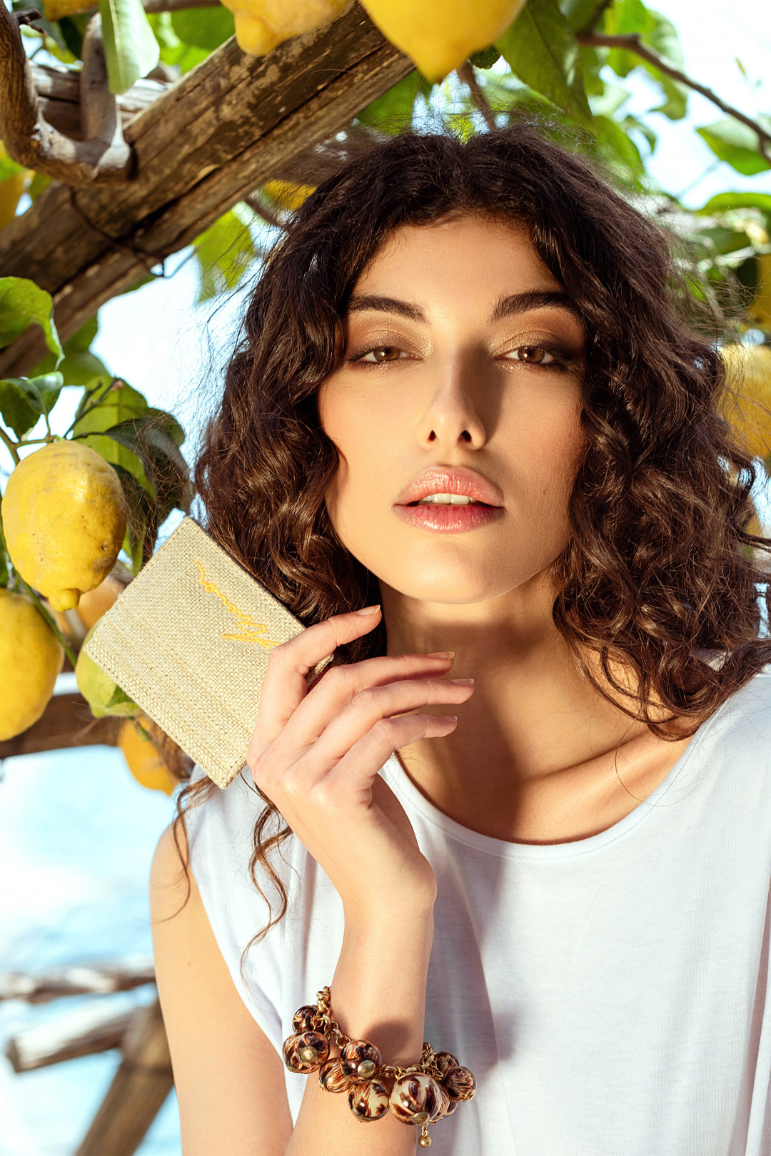 Woman holding a textured card, surrounded by lemon trees, wearing a white shirt and a decorative bracelet