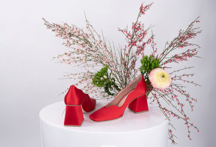 Red block-heel shoes displayed on a white pedestal with pink floral arrangement in the background