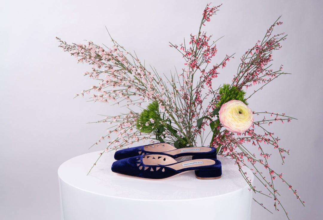 Blue velvet flat shoes displayed on a white pedestal with pink and white flowers in the background