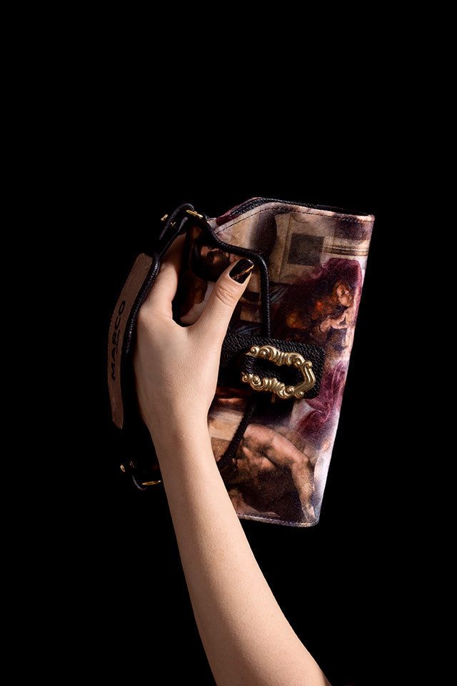 Hand holding a stylish designer clutch with classical art print and detailed clasp against a dark background