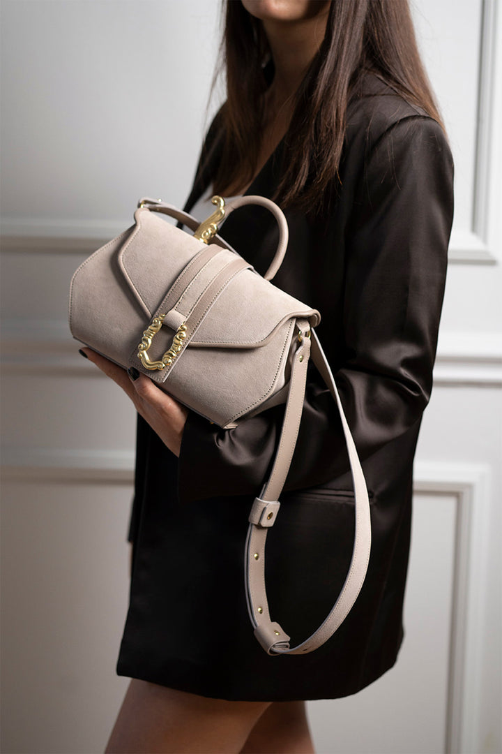 Woman holding a beige leather handbag with gold hardware against a white paneled wall