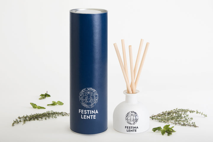 Blue cylindrical packaging and white reed diffuser with Festina Lente branding surrounded by fresh herbs