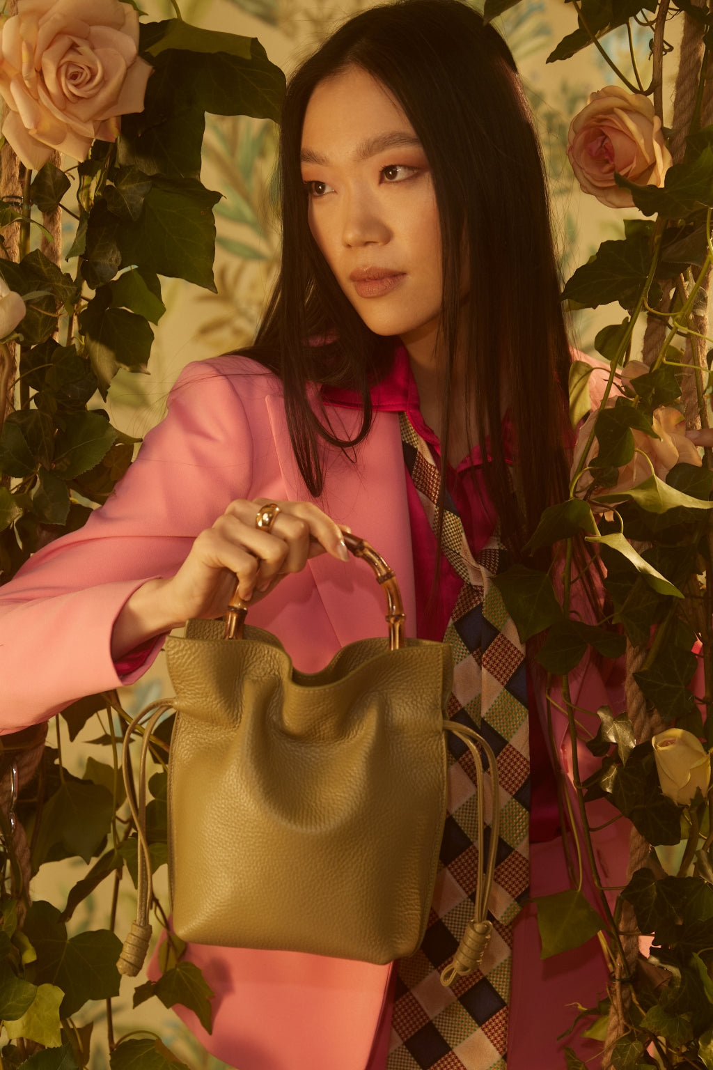 Woman in pink blazer holding stylish green handbag surrounded by flowers and greenery